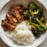 Top-down view of glazed chicken thigh recipes with rice and roasted broccoli in a rustic bowl on a marble surface.