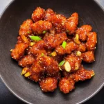 Top-down view of a dark bowl filled with a generous serving of Crispy Bang Bang Chicken Recipe with Spicy-Sweet Sauce, garnished with sesame seeds, green onions, and garlic slices on a dark, textured surface.