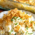 A vibrant photograph of a serving of Million Dollar Chicken Rice-A-Roni Casserole on a white plate, garnished with crispy fried onions and herbs, with a full baking dish blurred in the background.