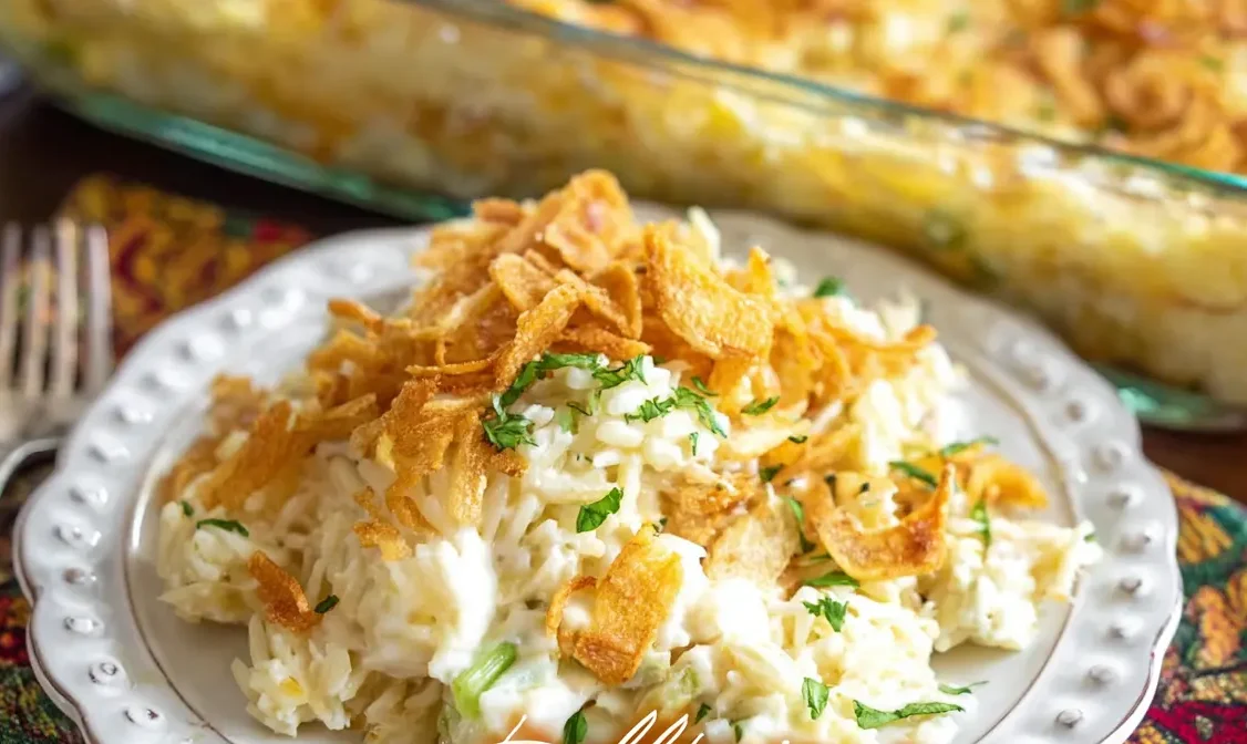 A vibrant photograph of a serving of Million Dollar Chicken Rice-A-Roni Casserole on a white plate, garnished with crispy fried onions and herbs, with a full baking dish blurred in the background.