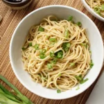 Overhead view of two bowls of savory garlic noodles garnished with fresh green onions, surrounded by whole garlic cloves and uncooked scallions on a rustic bamboo mat.