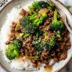 A rustic ceramic bowl filled with a delicious stir-fry of ground beef and broccoli over fluffy white rice, garnished with sesame seeds and scallions on a marble surface.