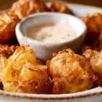 A close-up of golden-brown Bite-Sized Blooming Onions piled on a rustic plate with a creamy dipping sauce.