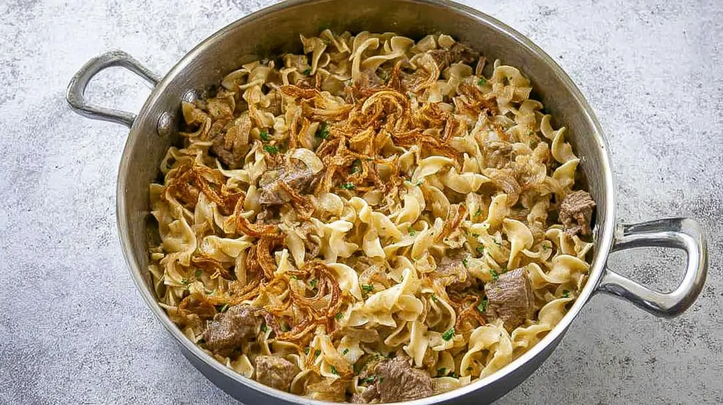 Hearty French Onion Beef and Noodles dish in a shiny metal pan, topped with crispy fried onions and fresh herbs.