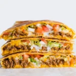 Stacked Keto Crunchwraps Recipe, cut in half to reveal seasoned meat, melted cheese, creamy sauce, fresh lettuce, and diced tomatoes on a white background.